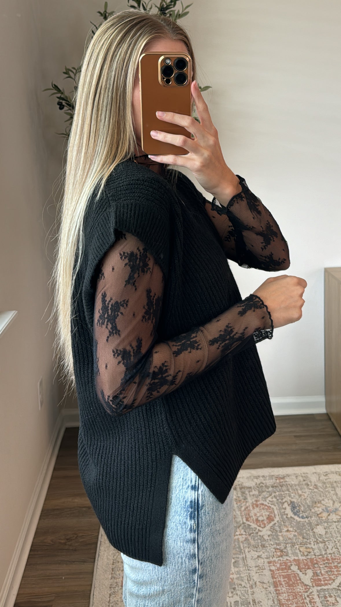 Person wearing a black sleeveless top with sheer sleeves and light blue jeans indoors.